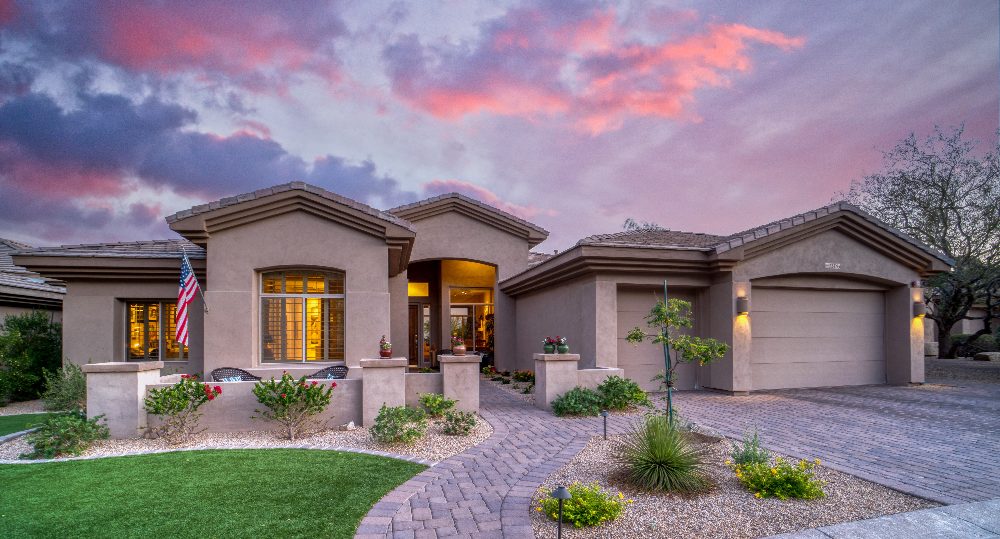 Modern single-story home at dusk with stucco exterior, arched windows, paver driveway, and desert landscaping