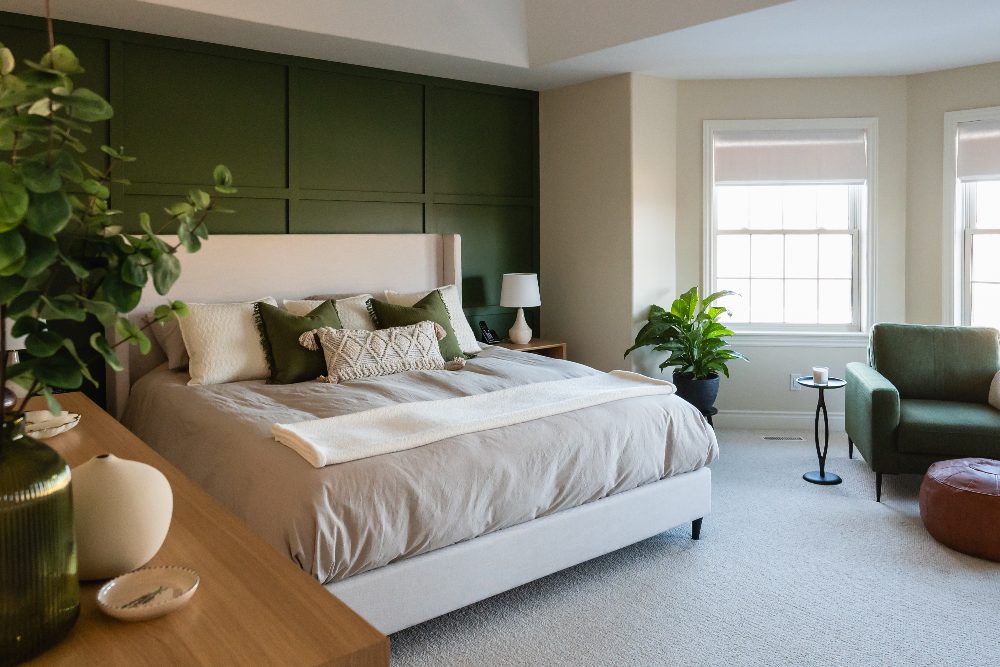 Modern bedroom with green accent wall, white bed, cream bedding, green chair, windows, potted plants, and wooden dresser